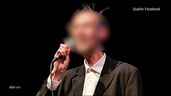 Ein Mann mit unkenntlich gemachtem Gesicht und lockerem Jackett steht mit einem Handmikrofon auf einer Bühne. © Screenshot 