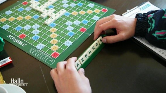 Ein Scrabble-Brett von oben. Spielsteine liegen bereits drauf. Davor sitzt eine Person, die mit zwei Händen die eigenen Spielsteine mit Buchstaben sortiert. © Screenshot 