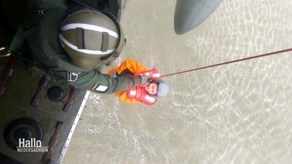 Eine Person wird an einem Seil aus dem Wasser in einen Helicompter gezogen. Die Person trägt eine Rettungsweste. © Screenshot 