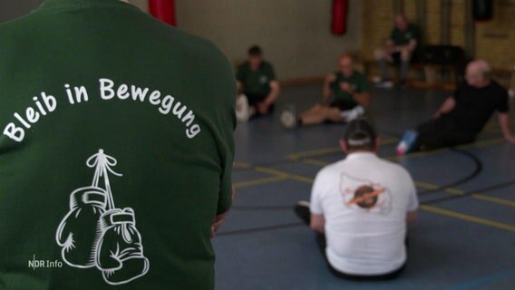 Ein grünes T-Shirt von hinten bedruckt mit der Aufschrift: "Bleib in Bewegung", darunter ein Logo von einem Paar Boxhandschuhe. © Screenshot 