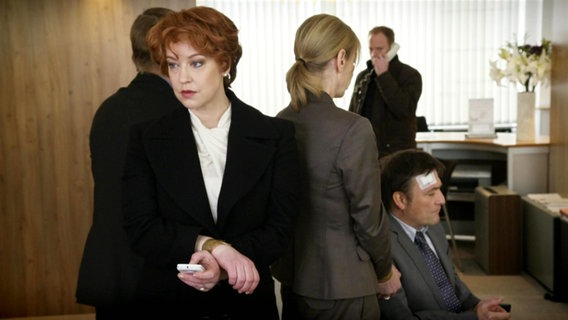 Drei Menschen stehen eng beeinander in einem Bürozimmer, ein Mann mit Kopfverletzung sitzt neben ihnen. Im Hintergrund telefoniert ein Mann. © Screenshot 