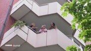 Drei Menschen stehen auf einem Balkon eines mehrstöckigen Hauses und unterhalten sich. © Screenshot 