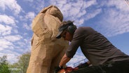 Ein Holzbildhauer arbeitet an einer Statue in Form eines bärtigen Mannes. © Screenshot 