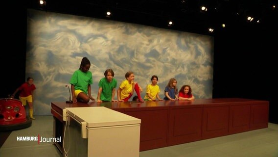 Mehrere Kinder klettern auf einen Schreibtisch auf einer Theaterbühne. © Screenshot 