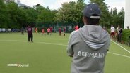 Ein junger Mann steht an einem Spielfeldrand und schaut anderen beim Hockeyspielen zu. © Screenshot 