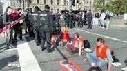 Mehrere junge Menschen sitzen auf einer Straße und blockieren den Verkehr. Vor ihnen steht Polizei. © Screenshot 