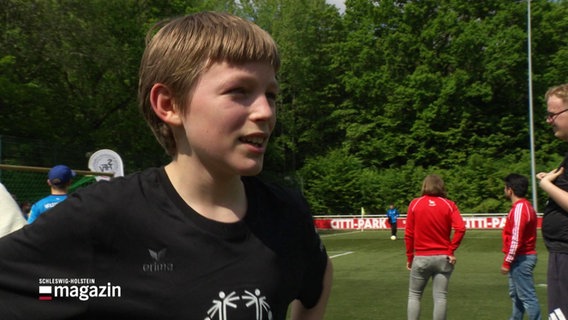 Ein Junge im Interview, im Hintergrund weitere Menschen auf dem Fußballplatz. © Screenshot 