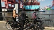 Drei Fahrradfahrerinnen gucken von der Hafenkante aus auf ein riesiges Container-Schiff, dass an einem Hamburger Container-Terminal vor Anker liegt. © Screenshot 