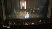 Szene in einer Oper: Eine Frau in weißem Kleid steht am Bühnenrand, ein Mann steht einige Meter neben ihr, auf der anderen Seite ein Chor von mehreren Personen. Im Orchestergraben dirigiert eine Dirigentin die Musizierenden. © Screenshot 