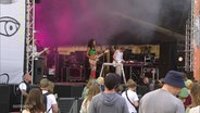 Auf einer Open-Air-Bühne bei einem Fetival spielt eine Sängerin mit bunten Klamotten und Gitarre um den Hals vor mehreren Menschen. © Screenshot 