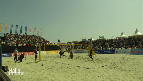 Blick auf eine Beachvolleyball-Arena an einem Strand in deren Mitte Spielerinnen bei einer Partie sind. © Screenshot 
