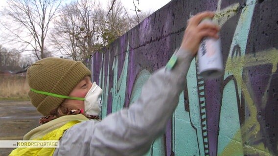 Eine Frau mit Atemschutzmaske malt Graffiti mit einer Sprühdose. © Screenshot 