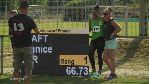 Zwei Athletinnen stehen Arm in Arm vor einer Tafel. Im Vordergrund ist ein Mann zu sehen. © Screenshot 