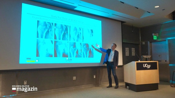 Ein Mann im Anzug steht in einem Hörsaal und deutet auf eine Leinwand auf die mehrere Röntgenbilder projiziert sind. © Screenshot 