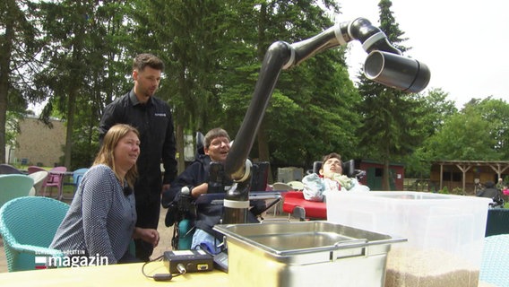 Eine Frau und ein Mann schauen einem Mann im Rollstuhl dabei zu, wie er einen auf einem Tisch stehenden Roboterarm bewegt. © Screenshot 