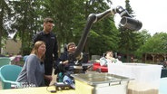 Eine Frau und ein Mann schauen einem Mann im Rollstuhl dabei zu, wie er einen auf einem Tisch stehenden Roboterarm bewegt. © Screenshot 