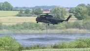 Ein Helikopter befüllt einen an ihm befestigten Behälter mit Wasser indem er knapp über einem See schwebt. © Screenshot 