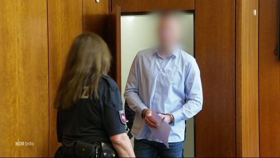 Ein Mann mit unkenntlich gemachtem Gesicht betritt, die Hände in Handschellen gefesselt, einen Gerichtssaal. © Screenshot 