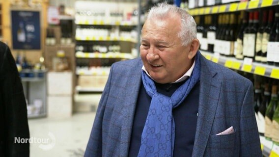 Ein älterer Herr mit Anzug und blauem Schal in einem Großmarkt. © Screenshot 