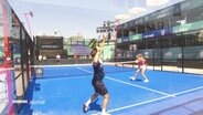 Ein Doppel-Spiel auf einem Padel-Feld. Ein Netz trennt wie beim Tennis das Spielfeld, um das Feld herum sind Begrenzungen, wie Plexiglasscheiben, die mit benutzt werden können. © Screenshot 