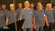 Mehrere ältere Herren mit blau-weiß gestreiften maritimen T-Shirts stehen laut singend als Shanty-Chor auf einer Bühne. © Screenshot 