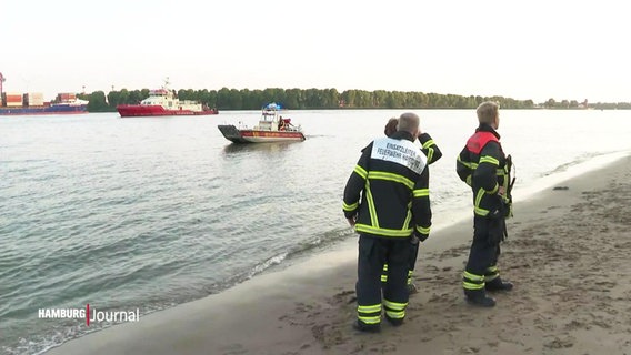 Rettungskräfte stehen in voller Montur am Elbstrand und schauen auf die Elbe raus. Auf dem Wasser selbst schwimmt ein Rettungsschiff. © Screenshot 