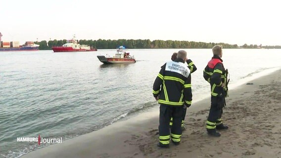 Rettungskräfte stehen in voller Montur am Elbstrand und schauen auf die Elbe raus. Auf dem Wasser selbst schwimmt ein Rettungsschiff. © Screenshot 