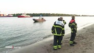 Rettungskräfte stehen in voller Montur am Elbstrand und schauen auf die Elbe raus. Auf dem Wasser selbst schwimmt ein Rettungsschiff. © Screenshot 