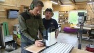 Moderator Arne-Torben Voigts bedient eine Holzfräse in einer Werkstadt, neben ihm steht ein Tischler begutachtend. © Screenshot 