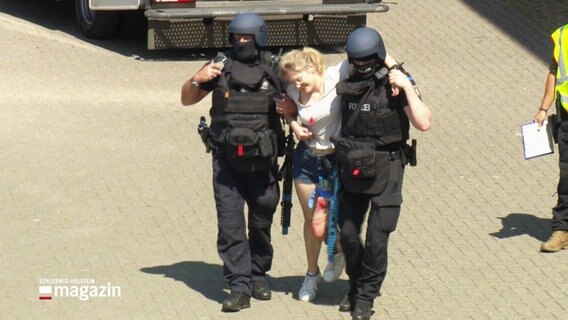 Zwei Polizisten in Sicherheitsrüstung stützen eine Frau, die eine Verletzte spielt und deren linkes Bein mit einem Gürtel abgeschnürt ist. © Screenshot 
