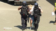 Zwei Polizisten in Sicherheitsrüstung stützen eine Frau, die eine Verletzte spielt und deren linkes Bein mit einem Gürtel abgeschnürt ist. © Screenshot 