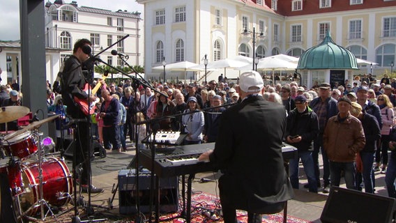 Bei einem Musik-Festival spielen Musizierende auf einer Bühne vor der viele Leute unter freiem Himmel tanzen. © Screenshot 