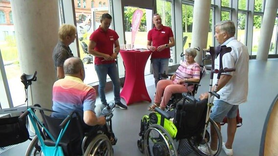 Mehrere Personen, zum Teil in Rollstühlen, hören zwei Männern in roten T-Shirts zu, die an einem Informationsstand etwas erzählen. © Screenshot 