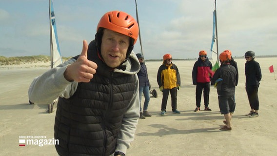 Schleswig-Holstein Magazin Moderator Henrik Hanses am Stranf von St. Peter-Ording. Er trägt einen Helm, schaut in die Kamera und macht einen Daumen nach oben. © Screenshot 