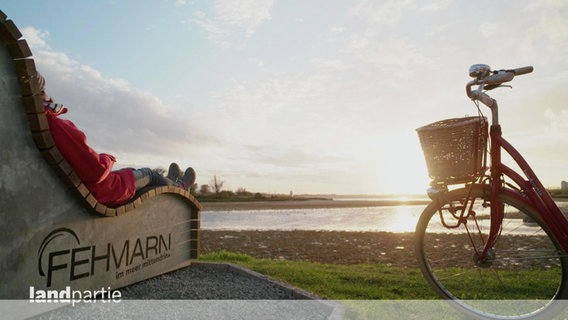 Eine Radfahrerin macht eine Pause auf einer Liege vor der Küste Fehmarns. © Screenshot 