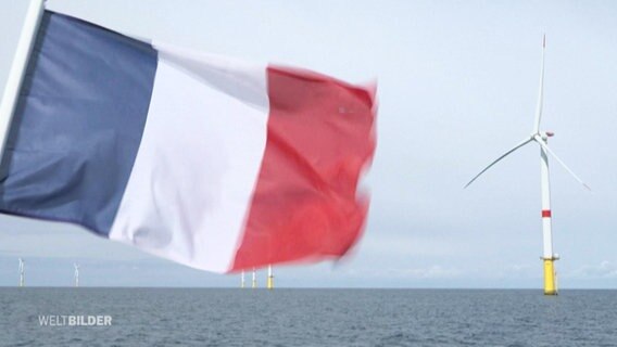 Die französische Flagge in Nahaufnahme weht im Vordergrund eines Windkraftwerks. © Screenshot 