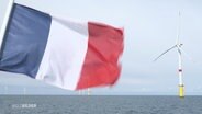 Die französische Flagge in Nahaufnahme weht im Vordergrund eines Windkraftwerks. © Screenshot 