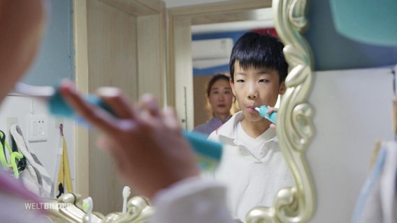 Ein Schulkind putzt sich die Zähne, während ihn seine Mutter durch den Spiegel beobachtet. © Screenshot 