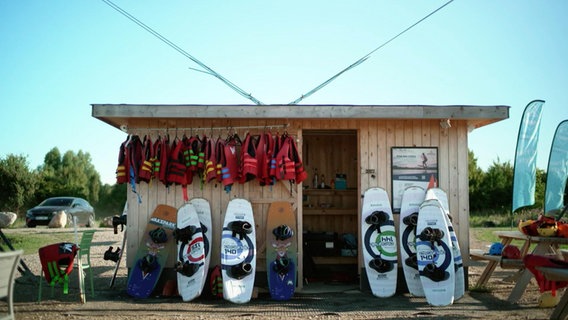 An einer Hütte lehnen mehrere Wakeboards und einige Rettungswesten hängen vom Dach. © Screenshot 