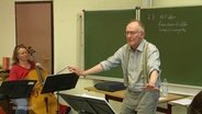 Ein älterer Herr dirigirt eine Gruppe von Musikern. © Screenshot 