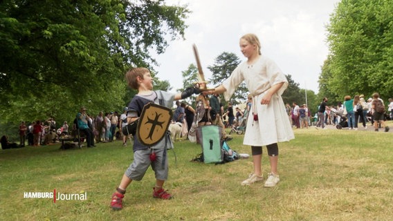 EIn kleinerer Junge und ein Mädchen kämpfen mit Schild und Holzschwertern auf einem Mittelalterfest in einem Park. © Screenshot 