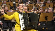 Ein Akkordeonspieler performt zusammen mit einem Orchester. © Screenshot 