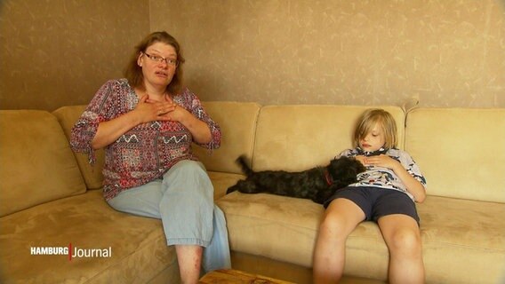 Frau sitzt links auf dem Sofa, mit beiden Händen auf dem Brustkorb. Rechts daneben sitzt ein Kind und in der Mitte des Sofas liegt ein Hund. © Screenshot 