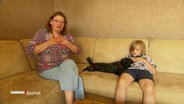 Frau sitzt links auf dem Sofa, mit beiden Händen auf dem Brustkorb. Rechts daneben sitzt ein Kind und in der Mitte des Sofas liegt ein Hund. © Screenshot 