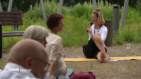 Frau sitzt auf einer Yogamatte. Das rechte Bein ist dabei über das linke Bein angewinkelt. Der Oberkörper rotiert aus der Hüfte heraus nach rechts.
Im Vordegrund sitzen zwei Damen und ein Herr, die die gleich Übung machen. © Screenshot 