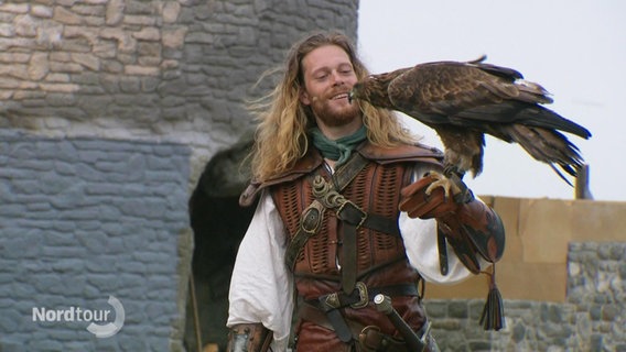 Ein Schauspieler mit langen Haaren und altertümlichen Haaren hält einen Adler auf seinem Arm. © Screenshot 