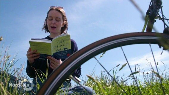 Eine junge Frau liest aus einem Buch. Sie sitzt im Gras, neben ihr steht ein Fahrrad. © Screenshot 