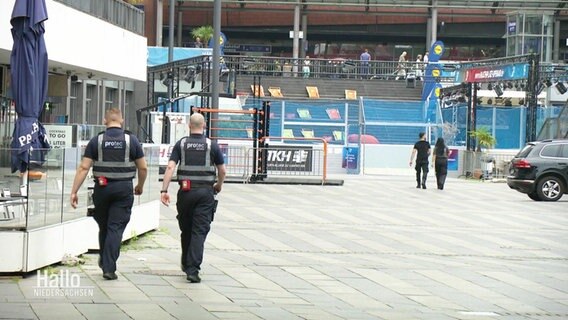 Zwei Security-Mitarbeiter gehen an einem öffentlichen Platz in einer Innenstadt entlang. © Screenshot 