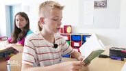 Ein Junge mit rot-weiß-gestreiftem T-Shirt sitzt in einem Klassenzimmer am Tisch und liest in einem Buch. © Screenshot 