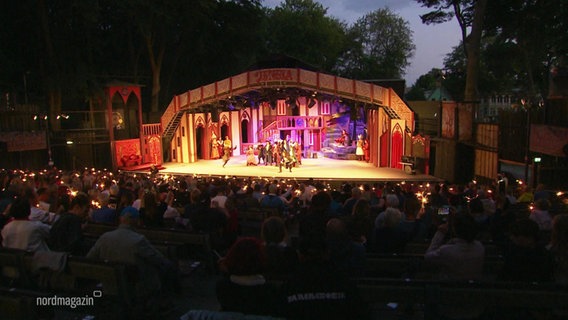 Auf einer Open-Air-Bühne führen bunt gekleidete Schauspielende ein Theaterstück vor gut besetztem Publikum auf. © Screenshot 